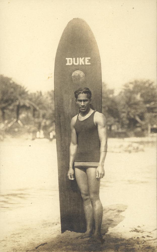 Duke Kahanamoku debout sur une plage devant sa grande planche de surf