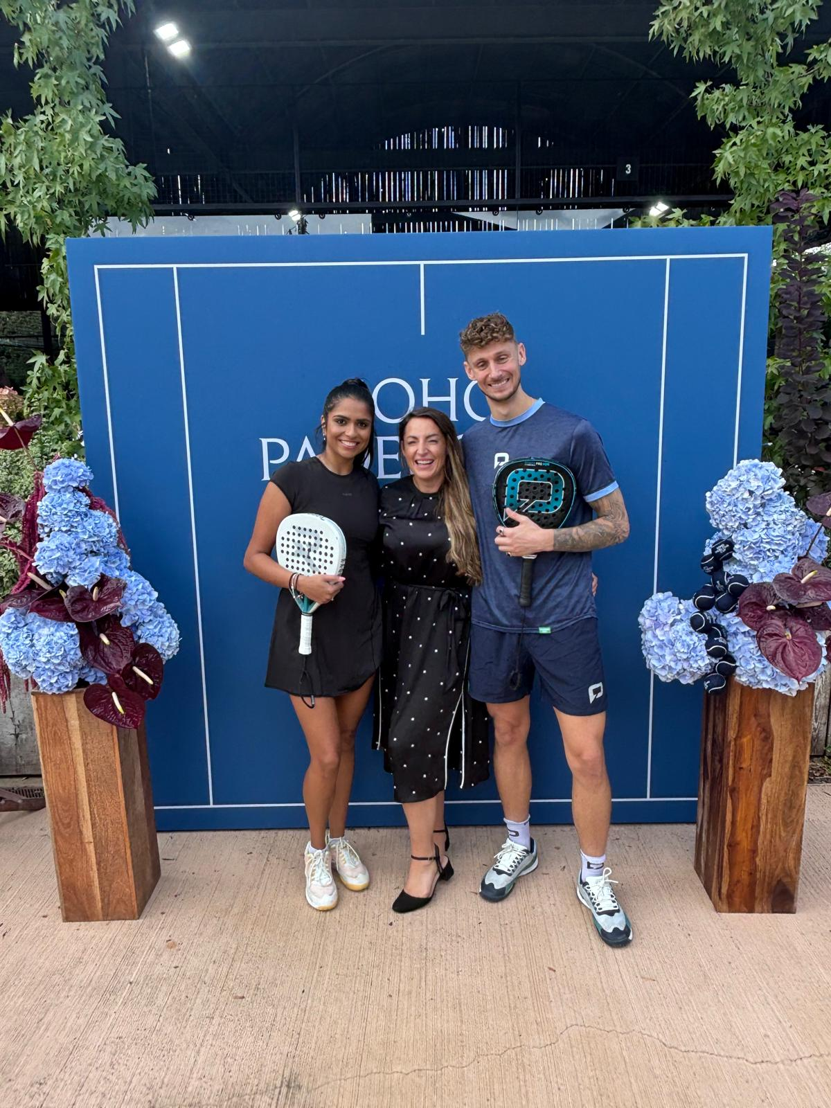 Aimée Lawson entourée de deux joueurs devant un décor bleu Soho Padel